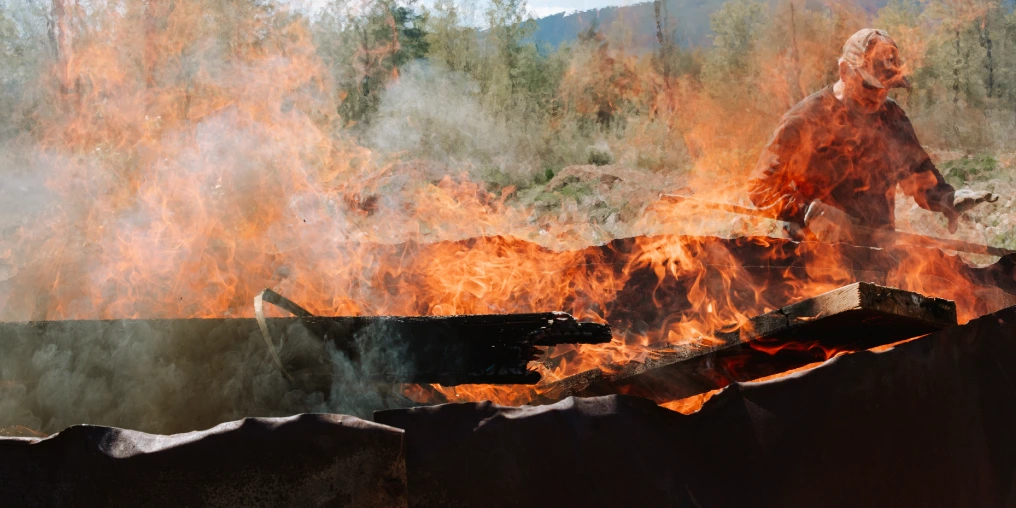 Person operating a flame cap biochar kiln in the field, adding woody biomass to create carbon sequestering biochar