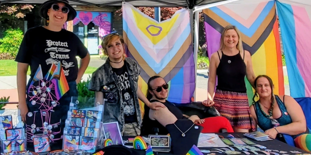 Members of Queer Centre Comox Valley at an outdoor Pride event booth displaying art and pride merchandise