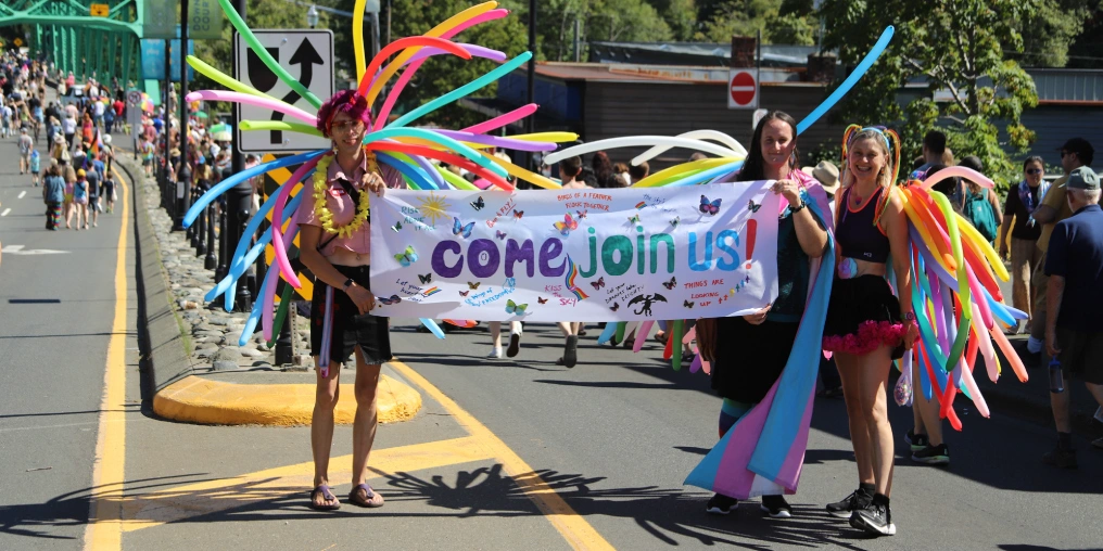 Participants in the Comox Valley Pride Parade marching across a bridge in Courtenay holding a Come Join Us banner