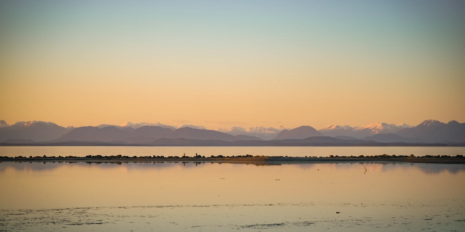 A golden sunset over calm water and a rocky sandbar in the Strathcona region, with snow-capped Coast Mountains in the distance, for Strathcona Collective Volume 13.