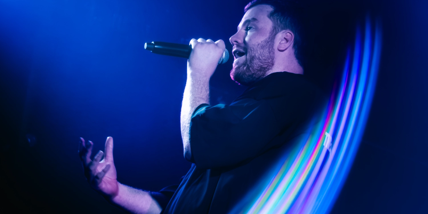 alick-mac-header Alick Mac performing on stage with microphone and blue stage lighting