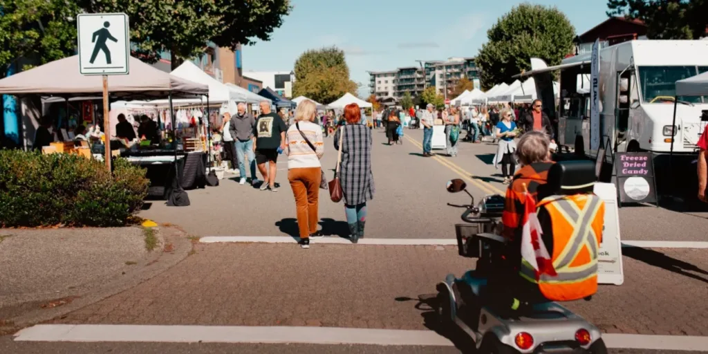 A wide view of the Campbell River Farmers Market on Shopper's Row, with vendors, shoppers, and a person on a mobility scooter in the foreground