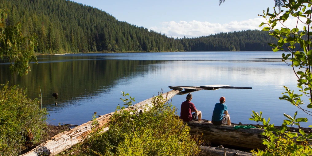 Two paddlers resting on a log overlooking a calm lake surrounded by forest on the Sayward Forest canoe routev