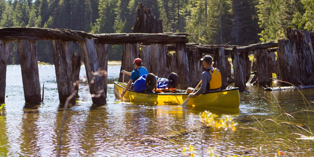 Two paddlers in a yellow canoe passing beneath the ruins of an old logging rail trestle on the Sayward Forest canoe route