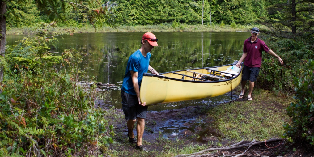 Two men carrying a yellow Clipper canoe through a muddy portage on the Sayward Forest canoe loop