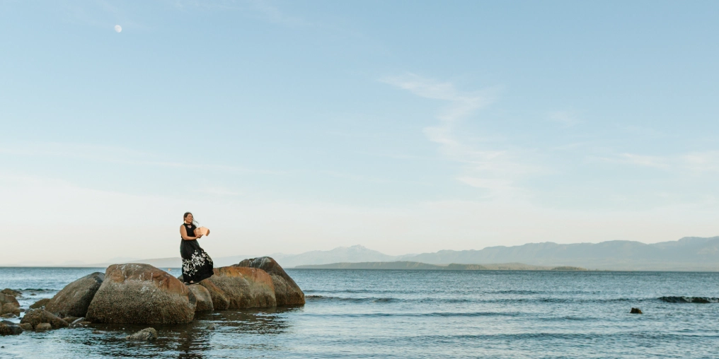 An Indigenous artist making waves in the Valley Agnes Seaweed Wisden stands on rocks along the Comox Valley coastline holding a copper art piece at dusk