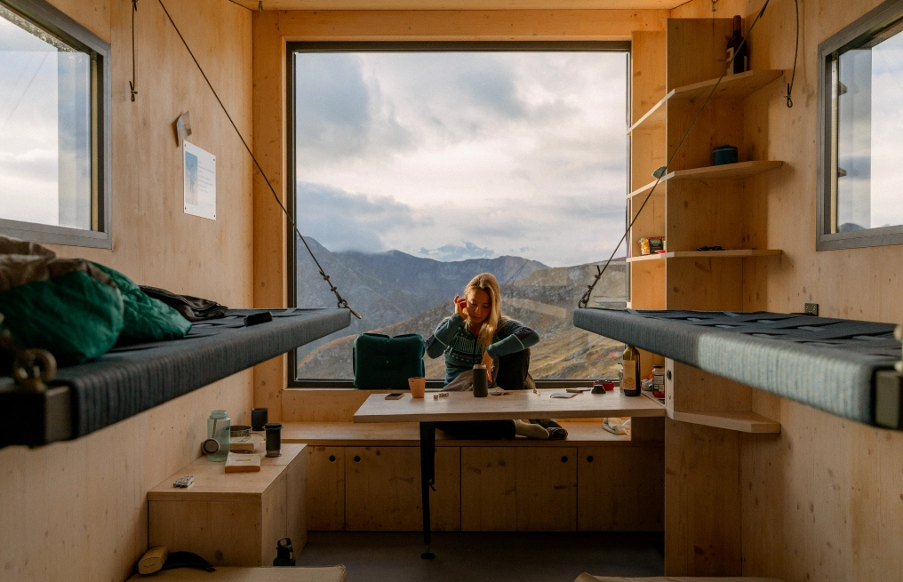 Interior of Bivacco Brédy with a hiker seated at the window table between two bunks