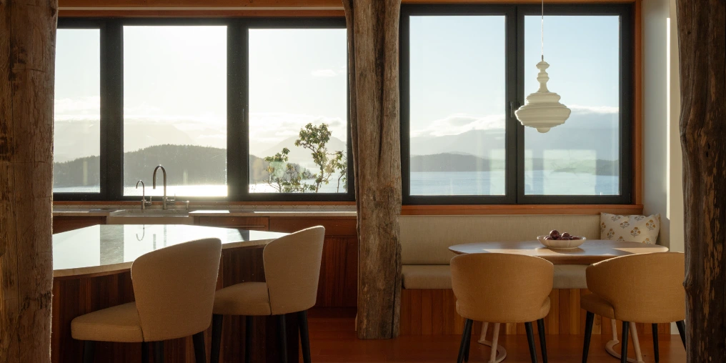 Capturing the perfect light at a special house on Cortes Island Wood-beamed dining nook with round table and built-in bench seating, framed by windows overlooking the water