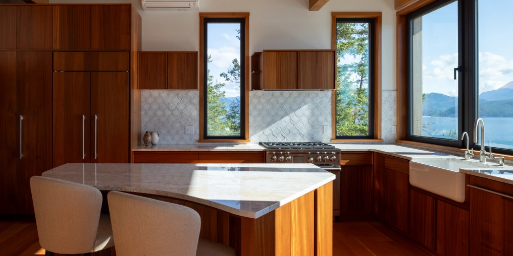 Capturing the perfect light at a special house on Cortes Island Kitchen with wood cabinetry and marble island, picture windows framing an ocean and mountain view