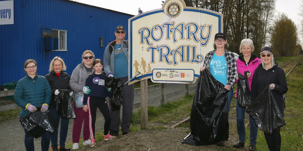 Celebrating 90 years of Rotary in the Comox Valley Volunteers holding garbage bags pose beside the Rotary Trail sign next to a railway line in the Comox Valley.