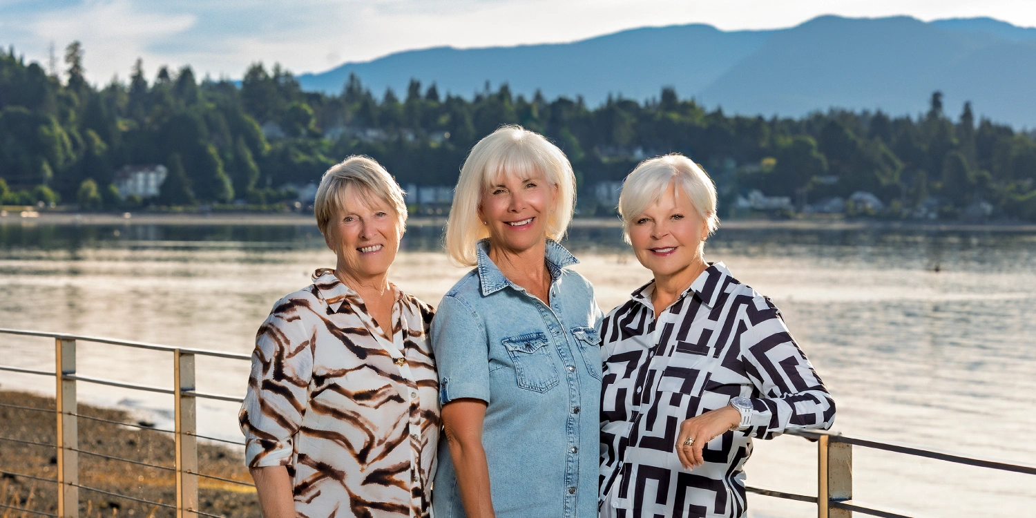Three women stand together at a railing by the water, with a forested shoreline and mountains behind them