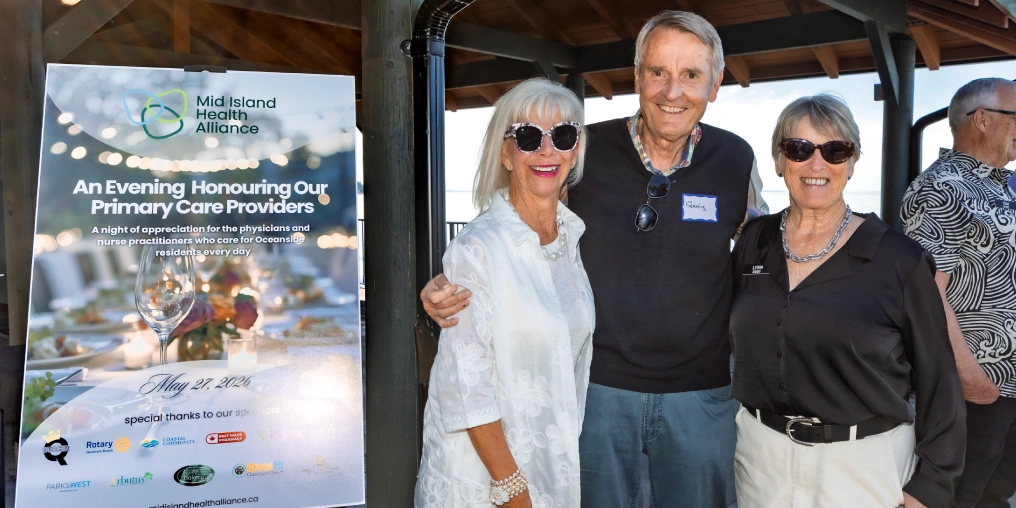 Three guests smile beside a Mid Island Health Alliance sign for An Evening Honouring Our Primary Care Providers, a May 2026 event for Oceanside physicians and nurse practitioners