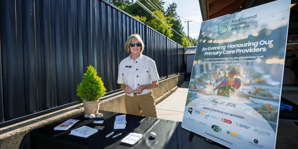 A woman stands at a welcome table with brochures beside a sign for the Mid Island Health Alliance evening honouring Oceanside primary care providers