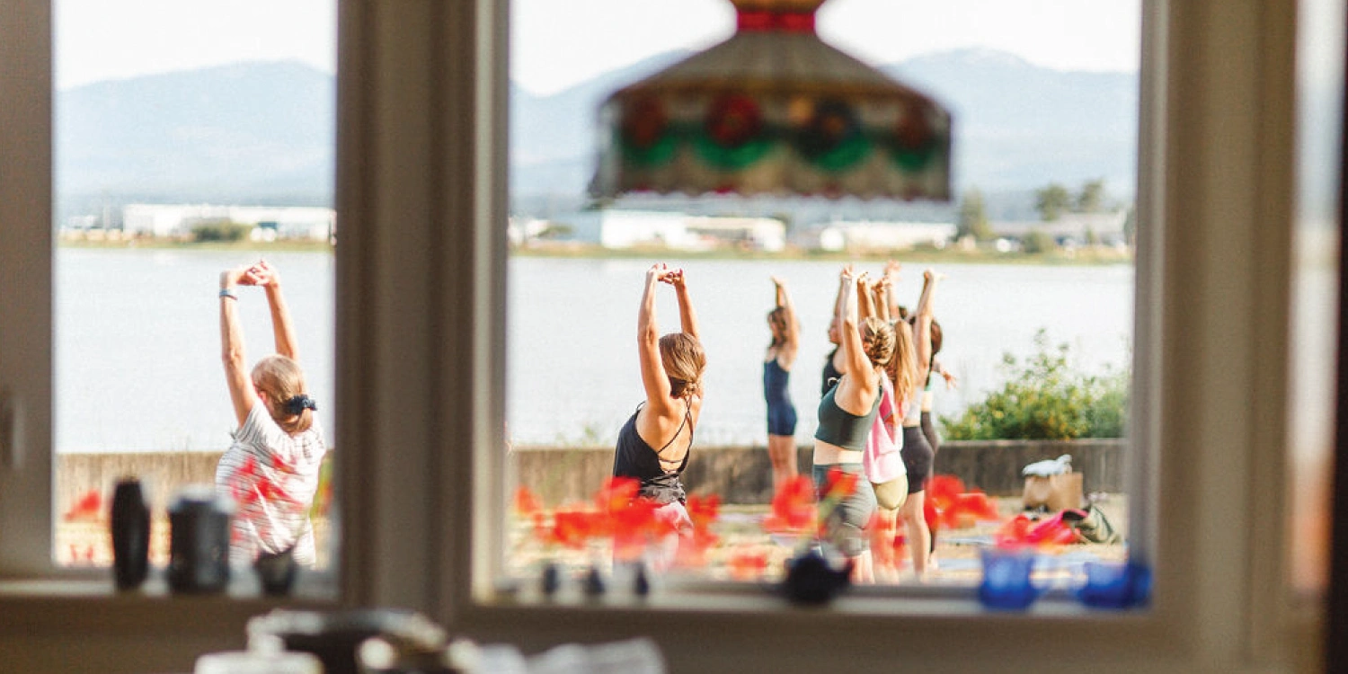 Yoga students stretching their arms overhead on a waterfront lawn, seen through a window framed by red flowers and a stained-glass lamp, with mountains across the water