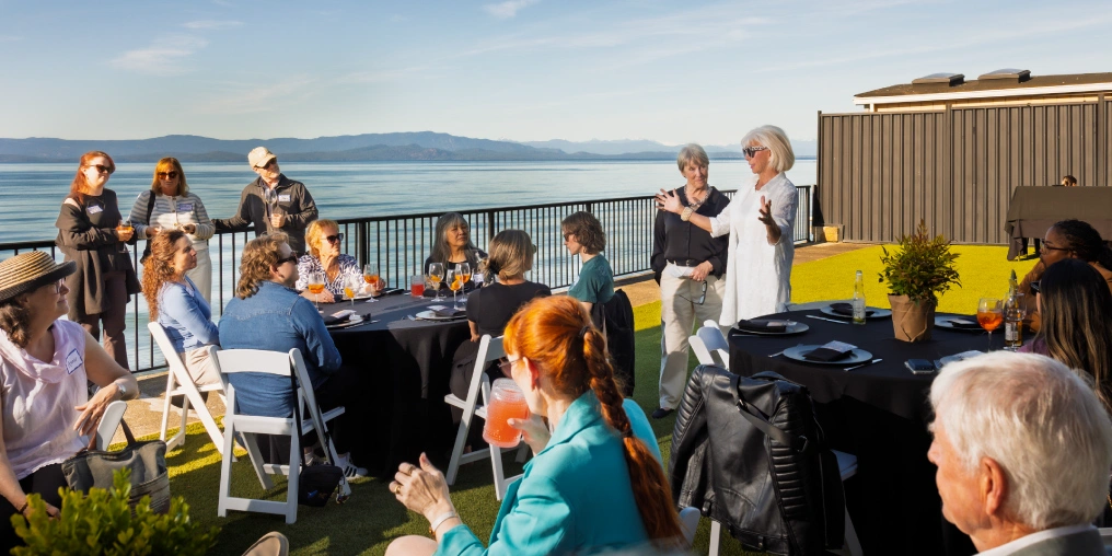 A woman speaks to guests seated at tables on an oceanfront terrace, with water and mountains in the background