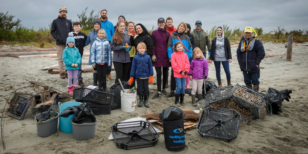 Celebrating 90 years of Rotary in the Comox Valley Adults and children gather on a sandy beach behind crates, buckets and wire cages collected during a shoreline cleanup.