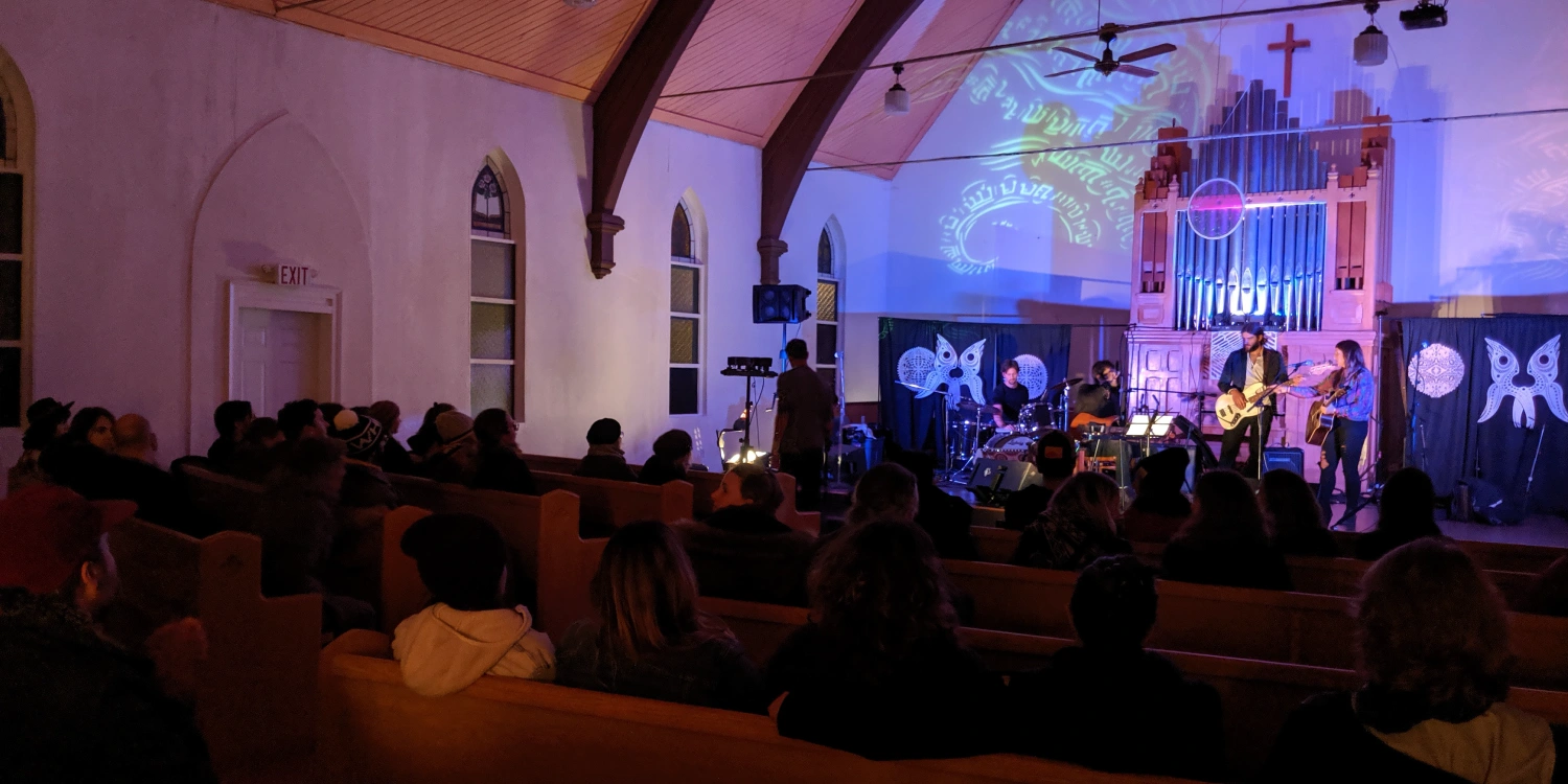 Band performing in front of the pipe organ at Weird Church as an audience watches from the wooden pews under projected lights