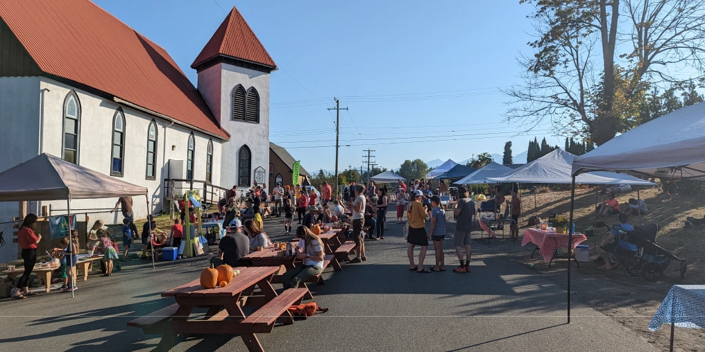 Outdoor community event beside Weird Church's white building and red roof, with pop-up tents, picnic tables and pumpkins along the street